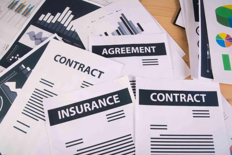 Top-down view of a disorganized pile of business documents on a wooden table, featuring papers labeled 'CONTRACT,' 'INSURANCE,' and 'AGREEMENT' mixed with data charts and graphs.