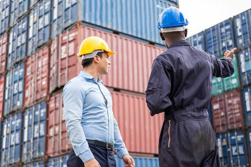 Two workers wearing hard hats standing in a large shipping yard surrounded by stacked colorful cargo containers; one worker in blue coveralls gestures towards the containers while the other in a light blue shirt listens.